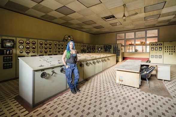 A woman with blue hair stands in an abandoned control room with antiquated equipment.