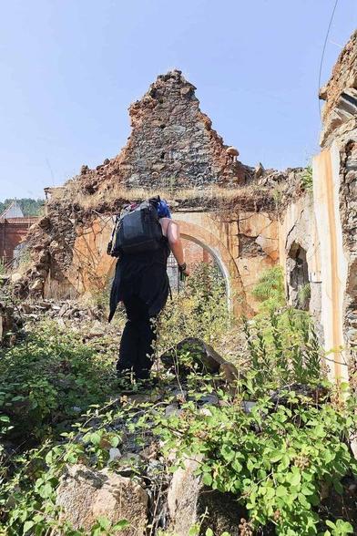 A backpacker explores the crumbling ruins of an abandoned building in Portugal. Overgrowth obscures the arched doorway of the structure, hinting at the decay found in abandoned places.