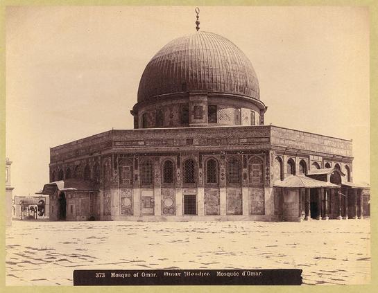 The image depicts a historical photograph in sepia tone, showing the "Mosque of Omar" also known as "Dome of the Rock" located in Jerusalem. The structure is an expansive domed building with intricate geometric patterns and Arabic inscriptions adorning its facade. It features multiple arched entrances and large windows that allow light to filter into the interior.

In front of the mosque, there appears to be a wide open courtyard paved with stones or bricks. A smaller secondary entrance can also be observed on one side. The photograph is labeled at the bottom as "373 Mosque of Omar," indicating its catalog number within a collection likely held by an archive like that mentioned in American Colony (Jerusalem).