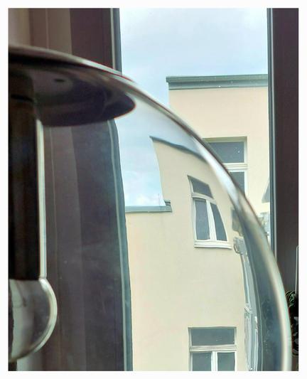View through a glass lamp from a room window onto a house opposite. The lamp, of which only the right half is visible, is a spherical ceiling lamp made of colourless glass, with a silvery bulb and dark socket. Due to the curvature of the glass, the house façade with its windows appears convexly curved and bent. Above the house is a bluish sky.