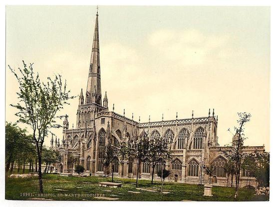 The image depicts an ornate Gothic-style church with intricate stonework, pointed arches, and a tall spire. This structure is identified as St Mary's Redcliffe in Bristol, England. The building features multiple levels of buttresses and windows that reflect the characteristic design elements of medieval English architecture. Surrounding the church are trees and grassy areas, suggesting it may be located within or near a cemetery, given some gravestones visible on the ground. The sky is overcast with soft lighting conditions, indicative of an outdoor scene during daylight hours without direct sunlight.