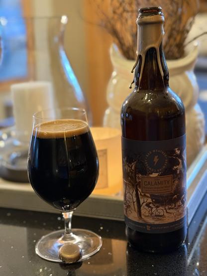 A tall snifter glass full of dark beer next to a 750ml brown bottle with a brown label and a gold wax neck label