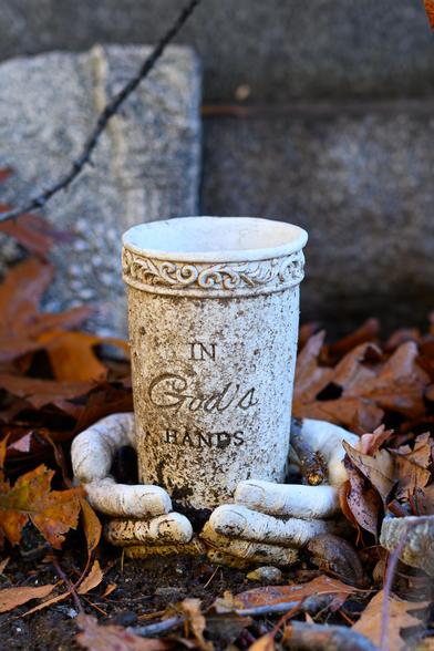 The photo shows a small decorative sculpture placed on the ground among dry brown autumn leaves. At the center is a weathered, off-white cup or vase made of stone or resin. It’s stained with age, giving it a rough, mottled texture. Around the rim is an ornate carved pattern of swirls.

On the front of the cup, dark engraved letters read: “In God’s hands.”

At the base of the cup, a pair of sculpted hands—also made of the same worn, pale material—appear to rise from the ground and gently cradle it. The hands look soft in their shape but are chipped and dirt-stained, as if they’ve been outdoors a long time. Fallen leaves and bits of soil gather around the fingers.

Behind the cup is a gray stone background that suggests this might be part of a memorial or gravesite. The overall mood is quiet and solemn, with the object looking lovingly placed but aged by time and the outdoors.