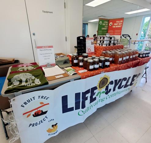 A community market table displaying jars of fig jam and apple juice, t-shirts, and hats, with signage advertising "Life Cycles" and "Fruit Tree Project", in an indoor space with white walls & glass doors on the right of the table. The wall beside the table, on the right, is mirrored.