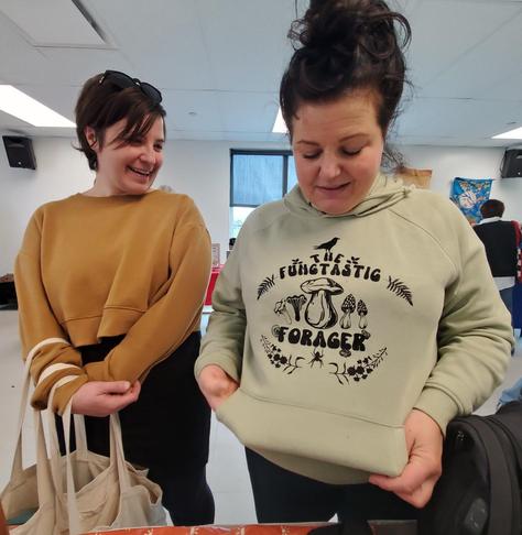 Two women are looking at a light green hoodie with the words "The Fungtastic Forager" printed on it, surrounded by illustrations of mushrooms, ferns, and a spider. The woman wearing the hoodie is holding it out to show off the design. The other woman, wearing a mustard yellow crop top, is smiling and holding a reusable tote bag. They are indoors with a white ceiling and walls and a red-patterned table in front of them.
