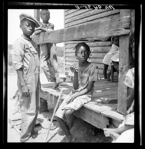 The photograph captures a group of young African American individuals, likely from the early to mid-1900s given its style and quality. It is black and white, portraying them in front of what appears to be an old wooden structure with vertical planks. Two boys are prominently featured; one on the left wears overalls with a hat and stands casually while looking away from the camera. The other boy sits on a bench or step holding onto a plank that's part of the building structure, his expression is more contemplative as he looks directly into the lens.

A girl in the center foreground holds an object near her mouth which could be interpreted as eating something; she wears shorts and has her hair styled with cornrows. Her gaze also meets the camera. On either side of this central figure are partial views of other individuals, suggesting a family or close-knit community setting. The condition of their clothes suggests modest economic circumstances.

The image is labeled "3-26-41" at the top, which could be indicative of its date when it was captured by Dorothea Lange for her work with photography documenting social issues and people's lives during that era in America.