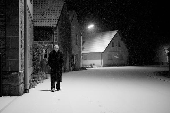 Dorf im dunklen. Straße entlang fotografiert. Die Straße biegt nach rechts ab. Links Häuser. Dunkel Beleuchtung. Ich gehe auf die Kamera zu. Mantel an und Jeans. Sneaker. Glatze. Brille.