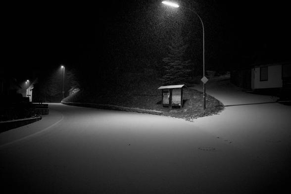 eine Straße biegt nach links ab. Die andere den Berg hoch nach rechts. Dorflampen entlang der Straße. Zwischen den Straßen sind Schilter. Vereinzelt Häuser. Dunkel. Beleuchtung.Straße Schnee bedeckt und es schneit.