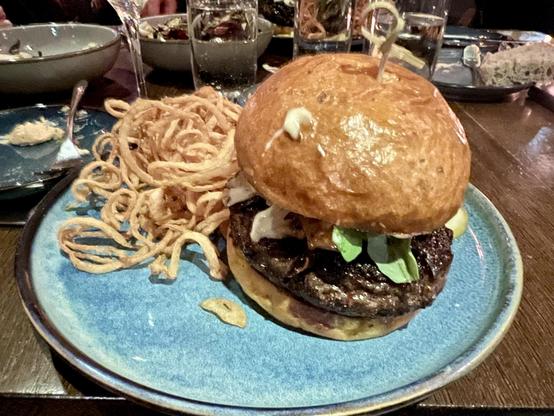 “SWEATA WEATHA” BURGER on blue plate on table. Bacon Shallot Jam, Maitake Mushroom, Dijonaise, Arugula,
“Fight For The Right” (Whale) Blue Cheese. Served with Crispy Onions. $24