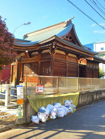Diagonal side view of the shrine and there is a yellow net over the adjacent garbage collection point full of bags.