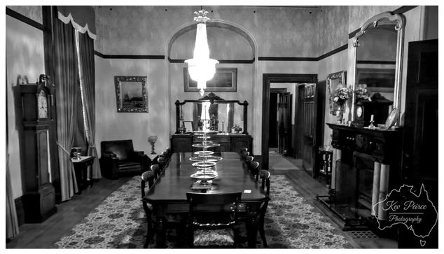 A wide, black and white photograph of the formal dining room at Martindale Hall. A long, dark wooden dining table dominates the center of the room, set with decorative plates and surrounded by matching chairs.  Above the table hangs a large, sparkling crystal chandelier, which is brightly lit and forms the central focal point under a high arched ceiling.  To the right is an elaborate fireplace and a tall, arched mirror reflecting the room. On the left stands a grandfather clock and heavy drapes.  The monochrome treatment emphasises the room's historical elegance and drama. The photograph is signed Kev Peirce.