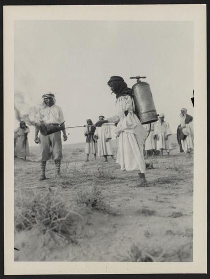 The image captures a group of people in an outdoor setting, likely from the early 20th century given its historical context. Two main figures stand out; one is dressed in traditional attire with a long robe and headscarf, carrying a large metal container on his back that resembles an early form of portable water tank or chemical dispenser. He holds another similar object in his hand which appears to be emitting smoke or vapor.

The second figure stands facing the first person, holding what seems to be a pipe-like instrument pointed towards the ground with one end glowing brightly. This suggests some sort of activity involving fire or flame control. The individuals around them wear loose-fitting clothing typical of desert-dwelling communities and appear attentive to the ongoing action.

In the background, more people can be seen observing from different distances. Their posture indicates interest but not active participation in what is happening between the two primary figures.

The scene takes place on barren ground with sparse vegetation, indicative of a dry environment such as a desert or steppe region. The photograph's monochrome tones and texture suggest it may have been taken using early photographic techniques like gelatin silver print.