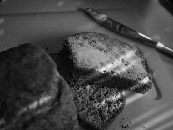 A thick slice of dark, rustic bread sits on a white cutting board with a generous pat of butter melting across the top. The butter glistens and pools in the bread's crevices, catching the soft light. More slices rest in the soft-focused foreground, their crumb catching the light. I butter knife rests above. The black and white treatment gives the scene a timeless feel, drawing attention to textures—the crusty bread, creamy butter, and grainy plate—rather than color.