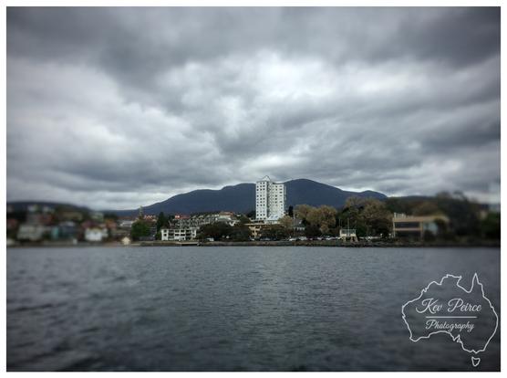 A photograph showing the approach to Hobart, Tasmania, with a focus on a tall, white building in the center. 
The foreground is a dark, slightly blurred body of water (the Derwent River).

The mid ground features the shoreline with low rise buildings and trees, and the background is dominated by the dark, majestic outline of Mount Wellington under a dramatic, cloudy sky.

The image has a tilt-shift effect, blurring the top and bottom.