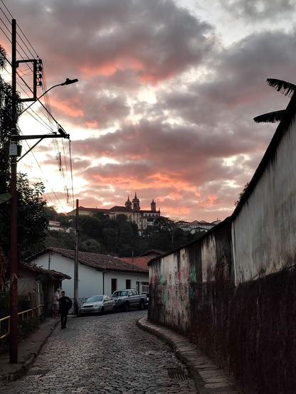 A dramatic sunset scene in Ouro Preto, Minas Gerais, Brazil, with an orange and pink sky reflecting on the clouds. A winding cobblestone street fills the foreground, with a person walking on the left side. On either side of the street are old, white-washed buildings and a stone wall adorned with graffiti. In the distance, silhouetted against the vibrant sky, sits a large, historic building complex atop a hill, likely the Igreja Matriz de Nossa Senhora do Pilar. Power lines crisscross the upper left corner of the frame.