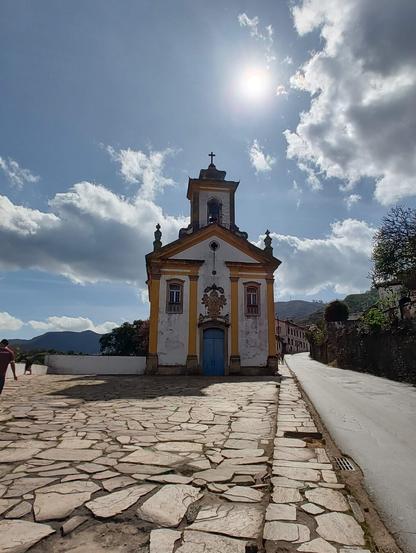 A low-angle shot on a sunny day captures the Oratorio do Padre Faria, a historic church in Ouro Preto, Minas Gerais, Brazil. The church features a white facade with yellow and orange trim, a blue door, and a bell tower topped with a cross. The cobbled stone ground of the town square stretches in front of the church, leading to a street on the right. In the background, under a partly cloudy sky with a bright sun, are mountains and distant buildings.