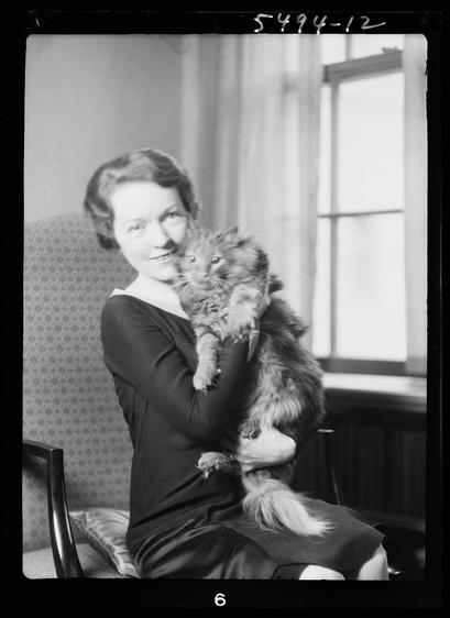 The image is a black and white photograph depicting an elderly woman sitting on a chair with her arms wrapped around two animals, likely pets. She has short hair styled in waves typical of earlier decades, possibly the 1920s or '30s, wearing what appears to be dark clothing.

In one arm she holds a large cat with thick fur and fluffy paws that suggest it may belong to a breed like Maine Coon known for its long coat. The other animal is smaller in size compared to the first, potentially another small pet such as a rabbit or rodent, held close against her body.

Behind them, there's an interior setting featuring curtains framing a window and patterned wallpaper on the wall. A number "5494-12" written at the top of the image suggests it might be part of a collection or cataloging system used by someone like Arnold Genthe known for his photographic work in that era.

There are no distinctive background elements that suggest location, likely to keep focus on the woman and her pets. A chair is visible beside them with a patterned design consistent with vintage interior decoration styles.