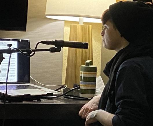 A person wearing a black beanie and scarf is sitting at a table with a laptop and microphone setup. A lamp and a striped container are visible in the background. The laptop screen displays text.