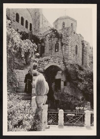 The image depicts a historic stone structure with multiple levels, featuring arches and arched windows. The architecture suggests it could be of historical or religious significance, possibly an old monastery or church building. There are several steps leading up to the main entrance, which is partially obscured by greenery. To the left side, there's a person in dark clothing standing at the top of the stairs, seemingly observing something below. At various levels along the path and structure, potted plants with spherical tops can be seen.
A group of people sits on steps near the bottom right corner, while others appear to be walking or sitting further back within the complex. The sky is overcast, giving a muted tone to the overall scene in this black-and-white photograph. There's no direct indication of modern technology such as cars, but it seems like an older setting based on attire and architecture style.
Information about traditional places of retirement for Zacharia suggests that the site might be associated with religious or historical significance related to early Christian monastic life.