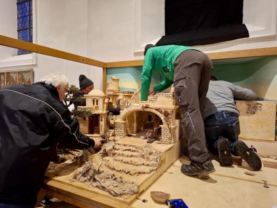 Auf einer großen Holzplatte wird eine Weihnachtskrippe aufgebaut: Zu sehen ist schon ein in Terrassen flach ansteigendes, felsiges Gelände mit trockenen Sträuchern und einem großen Baum. Dahinter werden Gebäude errichtet. Deutlich zu sehen sind ein breiter gemauerter Torbogen und ein verwinkeltes Gebäude mit Kuppeldach. Vier Leute sind mit dem Aufbau beschäftigt. Links steckt ein Mann mit grauen Haaren, Brille und dunkler Jacke Planzen ein. Von rechts beugen sich zwei Menschen in Jeans bzw. Arbeitshose, die direkt auf der Arbeitsplatte stehen, über die Stadt. Im Hintergrund arbeitet jemand mit grauer Mütze an den Rück- und Seitenwänden. Zu sehen ist auch, dass die Krippe eine gemalte Rückwand haben wird und gläserne Seitenwände. Das Ganze findet offenbar in einem Kirchenraum statt.