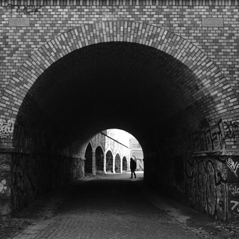 Das quadratische Schwarzweißfoto zeigt den Blick durch einen gemauerten Tunnel mit Rundbogen an Ein- und Ausgang. Hinter dem Ausgang ist links eine Wand mit 5 gemauerten Rundbögen, die die Form des Tunnels aufgreifen. Die Silhouette einer Person am Ausgang erscheint etwa an der Stelle, wo ein sechster Bogen sich anschließen könnte. Durch die leicht gebeugte Haltung wird die Ähnlichkeit zu den Bögen unterstützt. The square black-and-white photo shows the view through a brick tunnel with round arches at the entrance and exit. Behind the exit, on the left, is a wall with five brick arches that echo the shape of the tunnel. The silhouette of a person at the exit appears roughly where a sixth arch might join. The slightly bent posture reinforces the similarity to the arches.