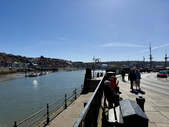 A riverside promenade runs alongside calm water with small boats moored near the bank, while a row of houses with red-tiled roofs rises on the opposite side. A paved walkway with benches and railings follows the curve of the river, where people are sitting and strolling in the sunlight. The road beside the walkway carries light traffic, including a bright pink car, and curves toward a harbour area with tall-masted ships. The sky is clear and blue, with a few faint contrails stretching across it, and the sun casts sharp shadows on the pavement.