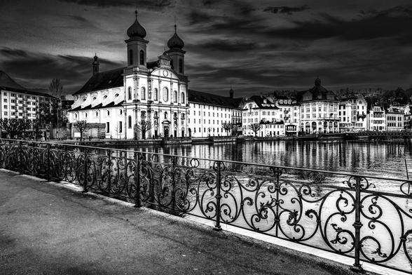 Jesuiten Kirche Luzern in Schwarz Weiss, von einer Brücke aus aufgenommen. Im Vordergrund ist ein Teil der Brücke mit einem verschnörkelten Geländer zu sehen. Die Kirche befindet sich im linken Drittel des Bildes, rechts davon sind mehrere Gebäude zu sehen. Das Licht der Gebäude spiegelt sich im Wasser.