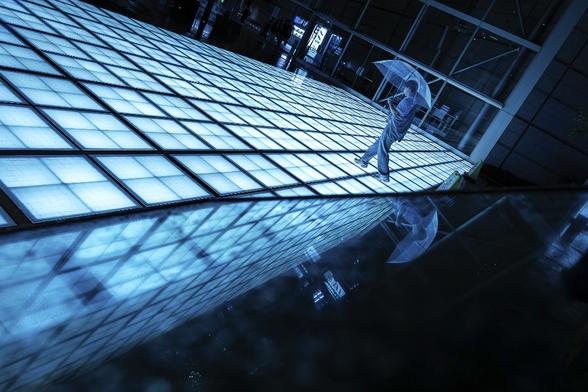 Reflection shot of a man with umbrella walking on a lit glass esplanade in Tokyo at night. The chosen angle leaves the impression that the man could fall of a flying structure. The scene is bathed in dark turquoise tones and surrounded by the night, leaving a cyberpunk atmosphere.