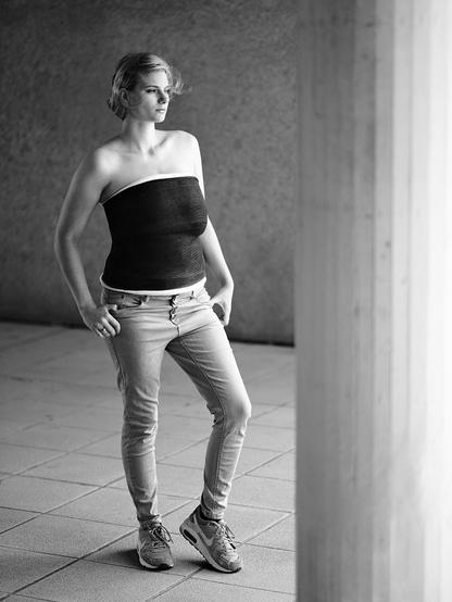 Eine junge Frau steht in einem hellen Raum mit großformatigen Plattenboden und einer Betonwand im Hintergrund. Sie trägt eine enge, schwarze Gipsmanschette, die ihren Oberkörper vom Brustkorb bis zur Taille umschließt. Die Frau hat kurze Haare und trägt eine helle, enge Jeans sowie Sportschuhe. Sie steht mit einer Hand in der Hosentasche und der anderen locker am Oberschenkel, leicht nach vorne geneigt.
