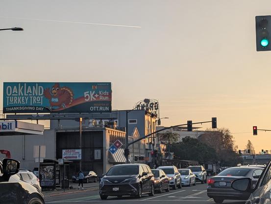 A late afternoon view of lanes of Grand Avenue traffic in Oakland, California with a sliver of the Grand Lake Theatre sign visible and a full view of the Oakland Turkey Trot 5K billboard