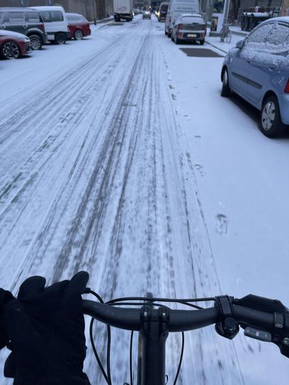 Eine Straße mit leichter Schneedecke und Spuren von Radfahrern im Schnee. Links und rechts am Straßenrand parken Autos die ebenfalls schneebedeckt sind.