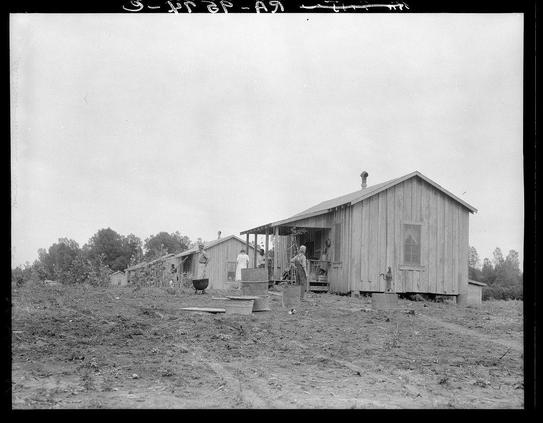 The black and white photograph depicts a rural setting with several wooden structures. The most prominent structure is a large, single-story building with visible weathering on the wood panels. To its left are smaller cabins or outbuildings also made of timber, suggesting an informal housing arrangement typical for workers' quarters during economic hardship periods such as in the Great Depression.

Several individuals can be seen around these buildings; one appears to be inside a cabin while others stand outside, possibly engaged in various activities that might include working on construction. The ground is unpaved and shows signs of wear with tracks from foot traffic or vehicles, indicating frequent activity in this area. A bare tree line borders the scene, hinting at sparse vegetation which could suggest an agricultural context.

Notably, there's a text overlay reading "3-6187," likely dating to 1937 given the year format and historical photography style associated with Dorothea Lange’s work during that time. The photograph is credited in smaller print as part of New cabins at Hill House, Mississippi by images.loener.nl.

The image conveys a sense of rural hardship, communal living or labor conditions prevalent before modern housing development became widespread. It also serves an educational purpose, potentially illustrating social history and the human condition during economic downturns like those experienced durin [...]