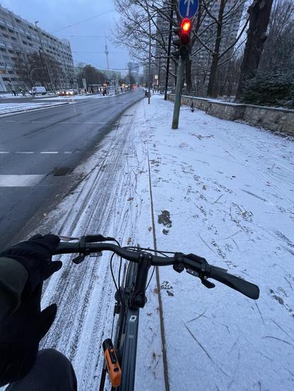 Eine Straße und ein Radweg. Der Radweg und der Fußweg rechts daneben sind mit einer leichten Schneedecke bedeckt. Der Radfahrer wartet an einer roten Ampel. Im Hintergrund Plattenbauten und der Berliner Fernsehturm