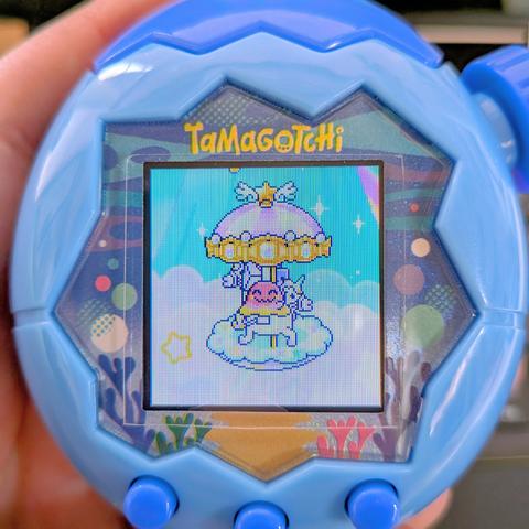 たまごっちパラダイスブルーの本体、まぐまっちが笑顔でメリーゴーランドに乗って遊んでいる。