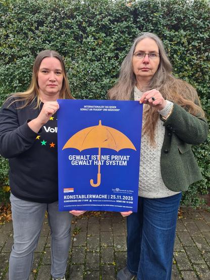 Zwei Frauen halten das Plakat mit dem Aufruf zur Kundgebung in die Kamera. Das Plakatmotiv ist ein gelber, aufgespannter Schirm, vor blauem Hinterrgrund. Der Schirm wirkt auf den ersten Blick solide, hat aber viele Löcher.
Die beiden Frauen, von denen einen Volt-Sweater trägt, wirken ernst und entschlossen.
