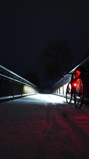 Ein beleuchtetes Gravelbike lehnt im Morgengrauen auf einer schneebedeckten Holzbrücke am Geländer.