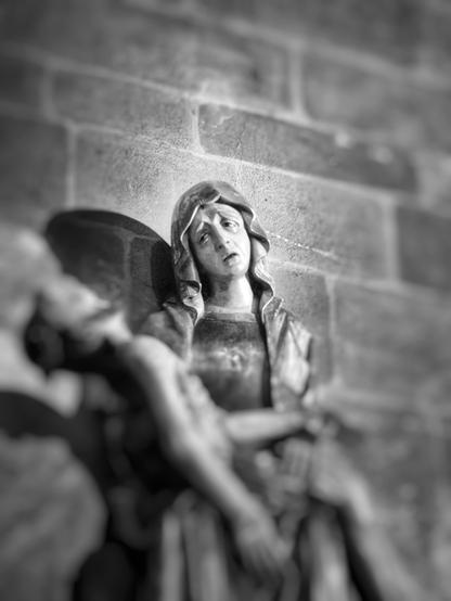 Schwarzweißaufnahme einer Frauenfigur mit verzweifeltem, schmerzerfülltem Gesicht. Der Kopf ist leicht zur linken Bildseite geneigt. Sie trägt ein Tuch auf dem Kopf, das bis über die Schultern fällt. Ihr Mund ist geöffnet und ihr Blick ist zur linken Bildseite gerichtet. Alles andere auf dem Bild, wie die gemauerte Wand hinter der Figur oder die männliche Figur, die sie in den Armen hält und auf ihrem Schoß liegt, ist unscharf.