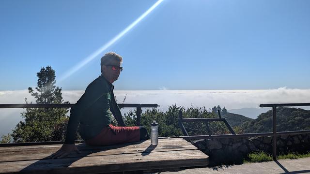 A man sits on a picnic bench in sun, near a mountain peak, high above a fog layer in the background