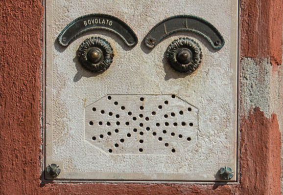 ein Klingelschild
nebeneinander zwei verzierte Klingelknöpfe (Augen), darüber je ein Bogen (Augenbrauen), auf dem linken steht der Name Bovolato. Darunter ein breit gezogenes Achteck mit vielen kleinen Löchern (Mund).