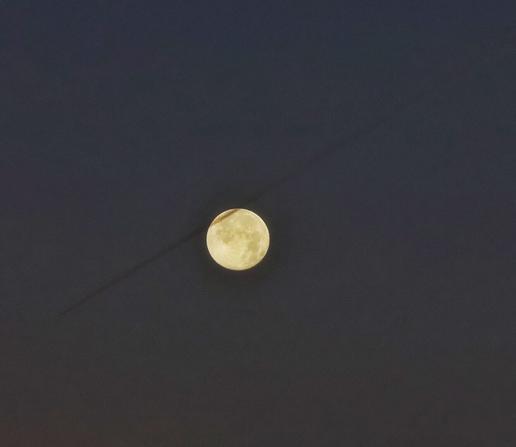 This image captures a tranquil early morning sky, featuring a luminous full moon just after the supermoon night. The moon, glowing with a soft yellowish hue, stands out against the deep, dark sky. A faint diagonal line, the contrail of an aeroplane, stretches across the lower portion of the image, adding a subtle contrast to the serene scene. The overall atmosphere is peaceful, showcasing the beauty of the moonlit sky in the quiet hours of dawn.