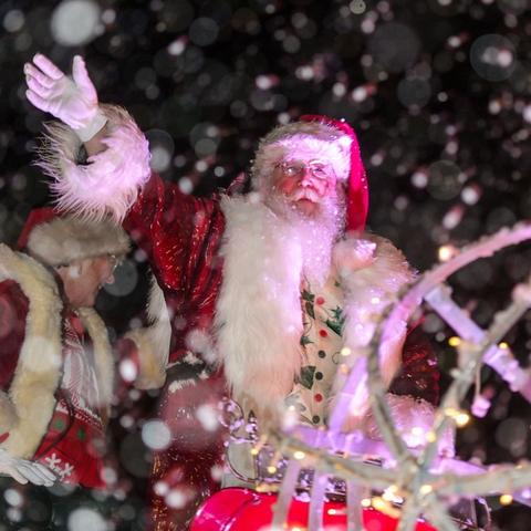 Santa Claus waving in snowfall, surrounded by lights and holiday decorations.