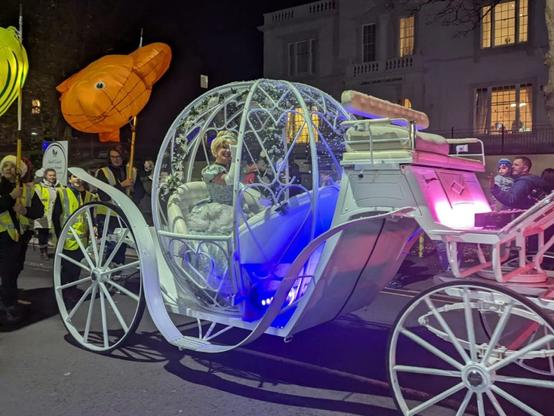 White illuminated Cinderella-style carriage with costumed performer inside, surrounded by fish lanterns.
