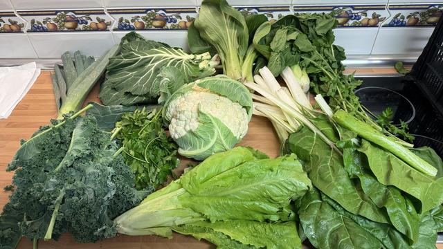 Contenido de la cesta de la semana sobre una encimera de cocina.
A la izquierda, desde abajo, kale y un puerro.
En el centro, una lechuga, perejil, una coliflor y pak choi.
A la derecha, acelgas, rúcula y más pak choi.