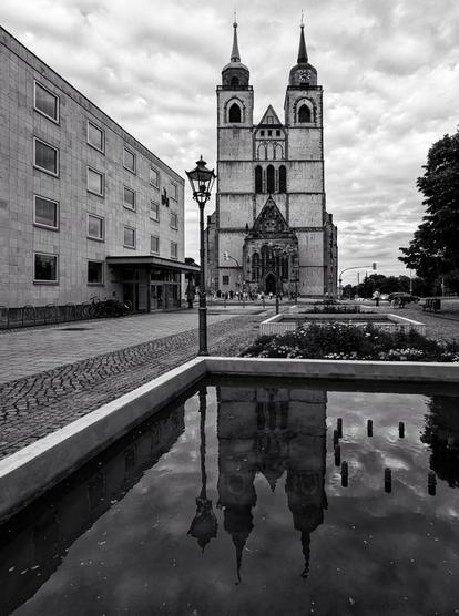 This black-and-white photograph features the Johanniskirche in Magdeburg, a historic church with two prominent towering spires. The church’s architecture is beautifully reflected in a still pool of water in the foreground, creating a mirror image.

To the left, a modern building with a minimalist design contrasts with the church’s ornate style, while a classic street lamp adds a touch of vintage charm. The cobblestone pavement and a small flower bed in front of the pool contribute to the timeless atmosphere. The overcast sky lends a moody, dramatic effect to the composition.