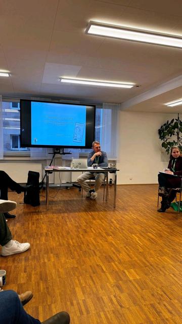Einblick in den Workshop: Die Teilnehmenden und der Referent Andreas Menne sitzen im Halbkreis vor der Präsentation zum Thema "Erinnerungskultur auf TikTok - Digital Memory in den medialen Lebenswelten von Jugendlichen" und diskutieren über Konsequenzen für die eigene Bildungsarbeit.