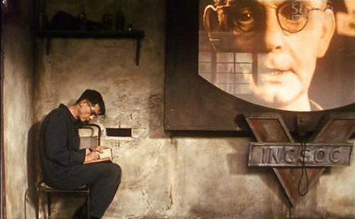 Fotograma de la película 1984 en el que se ve al protagonista sentado en un rincón donde no puede verle el gran hermano, que aparece en una telepantalla gigante que todo lo observa