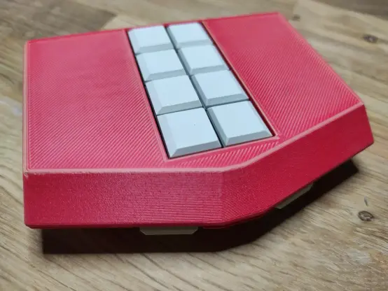 Mobile Braille-Tastatur Metabraille mit 8 Tasten auf der Vordreseite und zwei seitlich sichtbare Tasten auf der Oberseite. Das Gehäuse ist 3D-gedruckt, rot mit hellen Streifen und die Tasten sind weiß. Das Gehäuse ist ungefähr fünfeckig. Ein Rechteck wird an der Breitseite durch ein Trapez erweitert. Die Form ähnelt einem Haus mit Fenstern in der Mitte. Die Tastatur zeigt mit der Oberseite nach rechts unten und liegt auf einer Holzplatte.