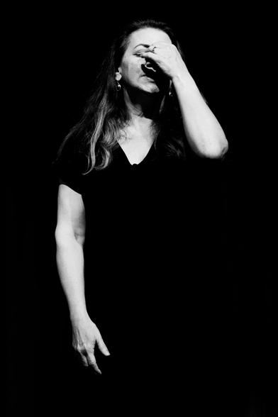 Against a completely black background, a woman stands. Her right arm hangs down, her left arm is raised towards her face, the fingers clasping the top of her nose. Her eyes are closed. She looks exasperated. Her dress is completely black and not visible.