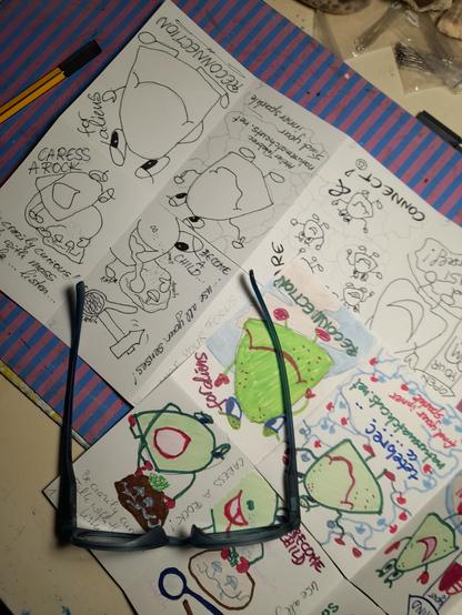 Two pages of paper with my glasses. One is provisional and roughly coloured, the other one is the fine drawing of the outlines in black, which I then colour in correctly. The main character is a friendly, chubby green alien with googly eyes, who represents Homo sapiens.