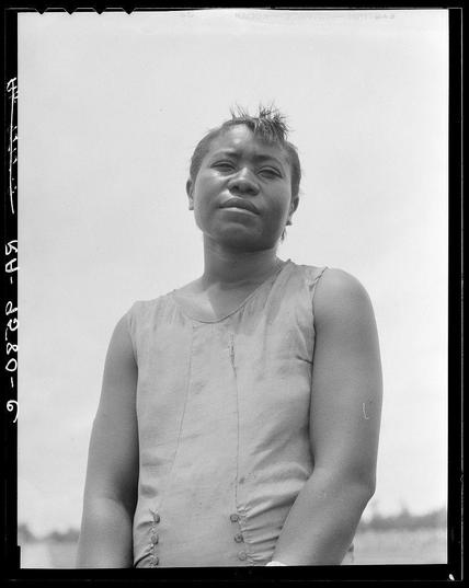 The photograph captures a woman standing outdoors with her gaze directed slightly to the side of the camera. She appears contemplative or pensive, and there is visible moisture on her skin which could be indicative of recent activity such as swimming or exercise. The black-and-white image highlights contrasts in texture and tone across different parts of her figure; particularly noticeable are the sheen on her hair, suggesting it may have been recently wet.

She wears a sleeveless dress with vertical button embellishments that add detail to its design. Her posture is relaxed but composed, as if posing for the photograph intentionally rather than captured spontaneously in action or movement.

The background is overexposed and lacks distinct features which suggests an open outdoor setting under bright sunlight. There are no discernible elements providing context about her location or surroundings beyond this being a broad daylight scene with clear skies.

There's textual information on the image, such as "DOROTHEA LANG" in the upper right corner likely indicating that Dorothea Lange was the photographer and possibly referencing her work from Mississippi Delta. The date '07-25-1938' suggests when this photograph may have been taken.

The overall mood of the image is somber, reflective or perhaps indicative of a labor-related setting given Lange's known focus on such subjects in historical documentation.
